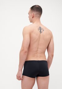 Schwarze Schwimmshorts aus elastischem Stoff mit glatter Textur und passgenauer Passform, Ansicht von hinten mit sichtbarem Tattoo im oberen Rückenbereich.