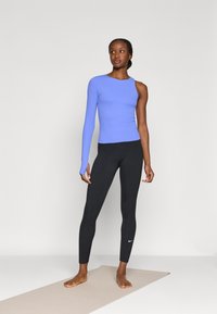 Ανοιχτό μπλε μακρυμάνικο τοπ με σχεδιασμό ενός ώμου σε συνδυασμό με μαύρο κολάν που φέρει λευκό λογότυπο Nike. Υφασμάτινη υφή, εφαρμοστή γραμμή.