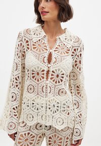 Crochet top in cream with floral patterns, long sleeves, and a pointed collar. Features button closure and sheer sections throughout.
