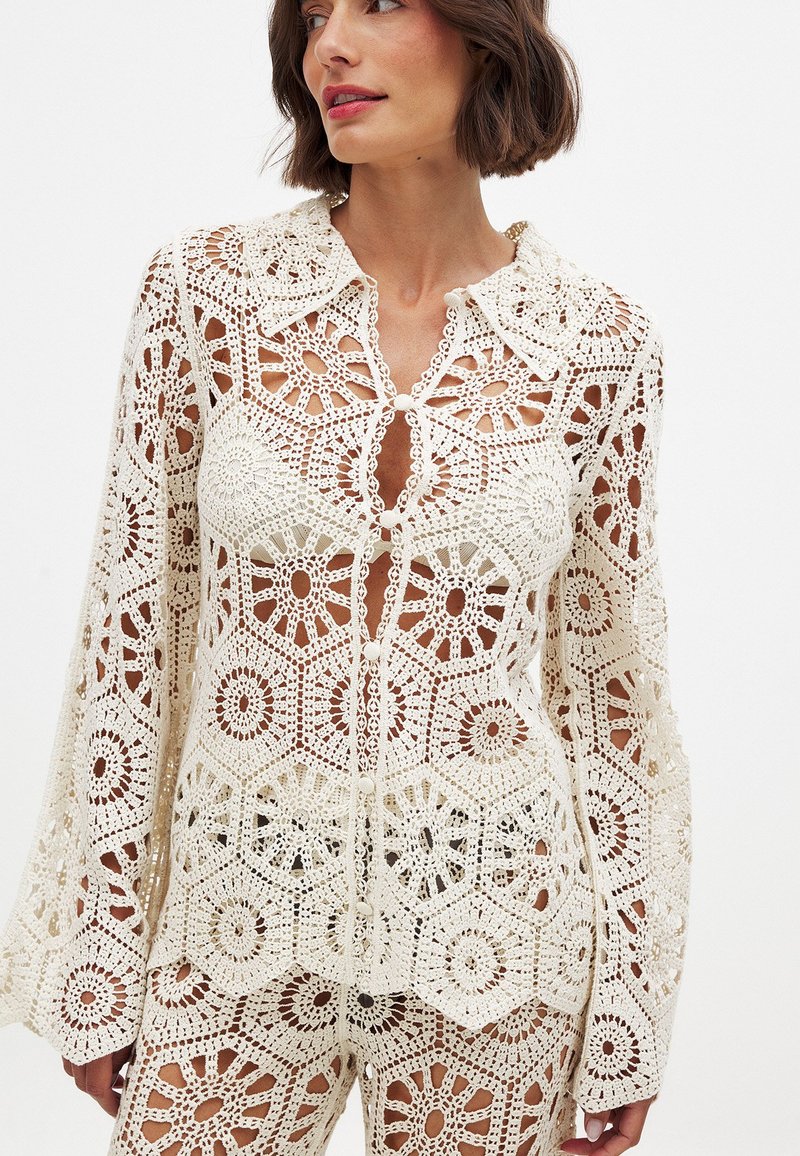 Crochet top in cream with floral patterns, long sleeves, and a pointed collar. Features button closure and sheer sections throughout.
