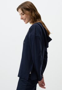 Felpa blu navy in tessuto morbido con vestibilità rilassata. Presenta un design con fessure laterali e lacci, abbinata a pantaloni scuri con texture.