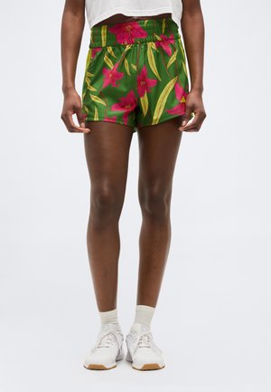 Persoon die groene short met roze bloemenprint draagt, een wit crop top, witte sokken en witte sneakers, staand tegen een effen achtergrond.