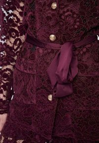 Indumento in pizzo bordeaux con intricati motivi floreali, bottoni dorati e cintura in raso coordinata annodata in vita. Sovrapposizioni testurizzate visibili.