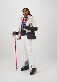 Moteris su slidinėjimo aprangoje, laikanti raudonus ir juodus slidinėjimo lazdas, vilkinti smėlio spalvos ir juodą striukę, baltas kelnes ir juodus slidinėjimo batus ant vientiso fono.