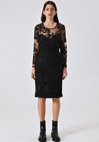 Robe en dentelle noire avec de longues manches transparentes, motif floral, silhouette ajustée et ourlet au genou ; associée à des bottines noires.