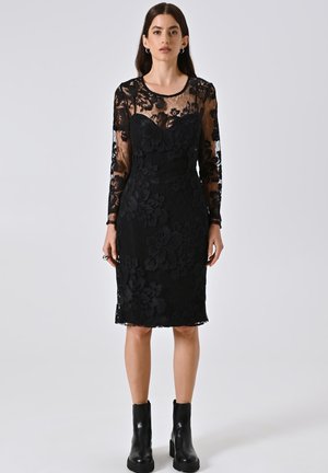 Schwarzes Spitzenkleid mit langen durchsichtigen Ärmeln, floralen Muster, figurbetonter Silhouette und knielangem Saum; kombiniert mit schwarzen Knöchelstiefeln.