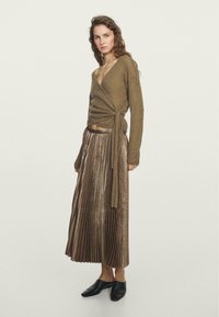 Pull enroulé marron à manches longues associé à une jupe plissée en bronze métallique. La tenue est complétée par des chaussures noires à enfiler.