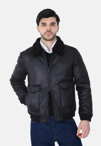 Uomo che indossa una giacca nera con collo in pelliccia, una camicia bianca, un maglione nero e jeans blu, in piedi con una mano in tasca contro uno sfondo bianco.