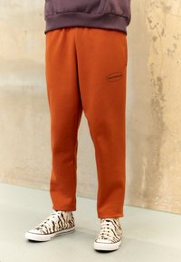 Oranga sweatshirtbyxor med elastisk midja och en svart "New Balance"-logotyp, tillsammans med svart och vit randiga sneakers.