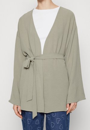 Vrouw draagt een licht olijfgroene wikkeljas met riem over een wit overhemd en blauwe broek met hartpatronen, getoond van nek tot heupen.