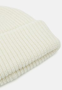 Valge kudumine beanie, millel on rööbastekstsuurus ja volditud säärenurk, valmistatud pehmest lõngast, mis näitab ühtlast mustrit kogu ulatuses.
