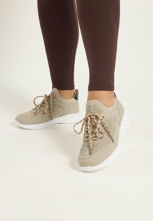 Person iført beige strikkede sneakers med hvide såler og mønstrede snørebånd, parret med mørkebrune tætsiddende bukser, stående på lyst gulv.