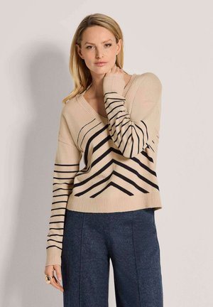 Blond vrouw draagt een beige V-hals trui met zwarte strepen en donkerblauwe broek, staat tegen een effen lichte achtergrond.