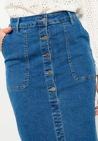 Denim krilo v srednji modri barvi, ki ima sprednjo gumbno zapiranje, dva stranska žepa in prilegajoč pas. Teksturna tkanina z diskretnimi šivi.