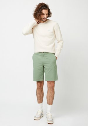 Homme aux cheveux bouclés portant une chemise crème à manches longues, un short vert sauge, des chaussettes blanches et des baskets, debout avec une main touchant son cou.