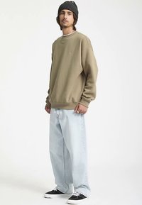 Sweat-shirt kaki avec poignets et ourlet côtelés, porté sur un jean ample bleu clair et des baskets noires, accompagné d'un bonnet noir.