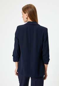 Blazer azul marino con cuello de solapa, mangas de tres cuartos y un diseño de espalda recta, hecho de tela suave, con bolsillos laterales.