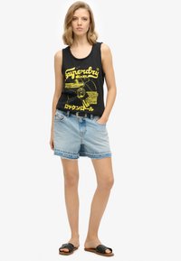 Top senza maniche nero con design grafico giallo, abbinato a pantaloni shorts in denim azzurro chiaro con orli sfilacciati e una cintura scura, indossato con sandali neri.