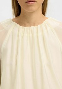 Kremfarvet bluse med en indsamlet halsudskæring, let gennemsigtigt stof og et løst, flydende design. Ingen synlige mønstre eller accenter.