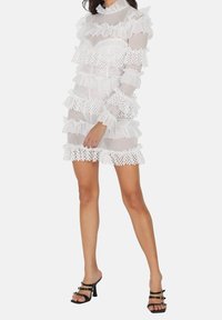 Robe blanche en voile avec des volants superposés et des accents en dentelle. Manches longues, col montant et une coupe fluide. Associée à des escarpins noirs à lanières.