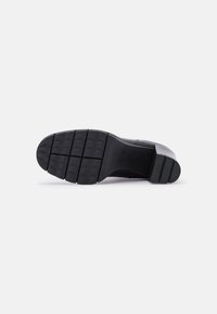 Semelle de chaussure noire en caoutchouc texturé, avec des rainures horizontales et une pointe arrondie. Partie supérieure lisse avec une légère plateforme.