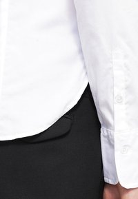 Camicia bianca a maniche lunghe con polsini abbottonati, infilata in pantaloni neri in tessuto con tasca laterale. Texture liscia, design classico del colletto.