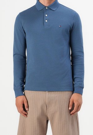 Mann trägt ein blaues langärmeliges Poloshirt und beige gerippte Hose, steht vor einem einfarbigen weißen Hintergrund.