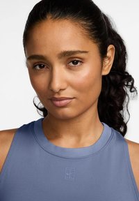 Lichtblauw sporttop met ribbeltextuur, voorzien van een ronde halslijn en subtiele logodetail. Model heeft donker, krullend haar.