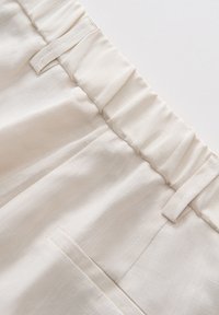 Crèmekleurige stof met een zachte textuur, voorzien van een geplooide tailleband en zijzak, ontworpen voor comfort en gemakkelijke draagbaarheid.