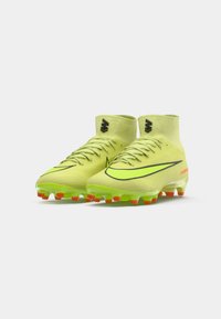 Un par de botas de fútbol de caña alta en neón amarillo-verde con logotipos negros y suelas con tachuelas naranjas sobre fondo blanco.