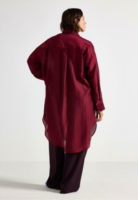 Žena s otočeným zády, oblečená v dlouhé burgundské košili s sklady a tmavých širokých kalhotech, stojí proti jednodušenému světlému pozadí.
