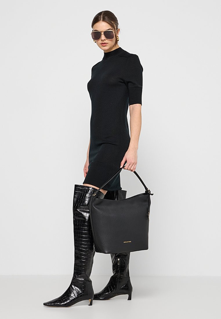 Borsa a mano in pelle nera con superficie liscia e una sola tracolla, abbinata a un abito nero aderente e stivali lucidi fino al ginocchio.