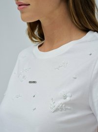 Camiseta blanca de algodón con bordado floral texturizado y cuentas. Cuello redondeado con un pequeño detalle de logo metálico en el centro.