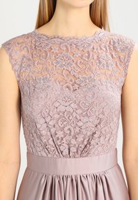 Robe en dentelle de couleur mauve doux, ornée de motifs floraux raffinés, avec des manches courtes et une ceinture en satin accompagnée de détails de jupe froncés.