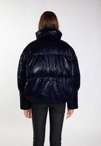 Marineblau gesteppte Jacke mit glänzender Oberfläche, hohem Kragen und gerafften Bereichen. Getragen über schwarzen, figurbetonten Hosen, die eine abgerundete Silhouette zeigen.