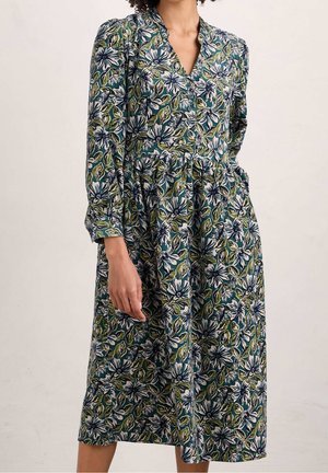 Vrouw draagt een halflange jurk met lange mouwen en een groen, wit en blauw bloemenpatroon, staand tegen een effen lichte achtergrond.