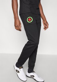 Čierne teplákové nohavice s okrúhlym zeleným nášivkovým prvkom s červenou hviezdou a textom. Model má na sebe biele športové topánky s čiernymi detailmi.