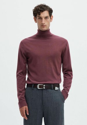 Homme portant un pull à col roulé bordeaux, un pantalon gris et une ceinture marron foncé, debout avec une main dans la poche sur fond blanc.