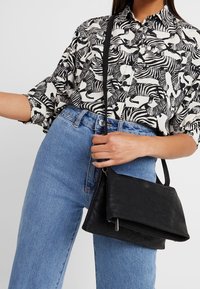Borsa a tracolla in pelle nera con design a patta ripiegata e accessori in argento, abbinata a una camicia a fantasia zebrate in bianco e nero e jeans blu.
