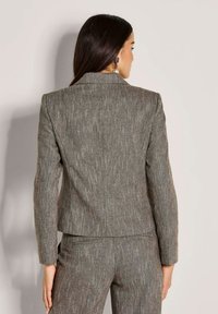 Vrouw met lang, zwart haar, gekleed in een grijze, gestructureerde blazer en bijpassende broek, staand tegen een vlakke, lichte muur, met de rug naar de kijker.
