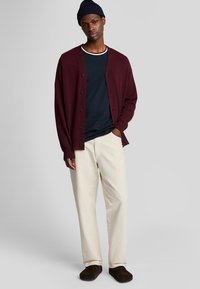 Cardigan bordeaux con bottoni sopra una camicia blu navy, abbinato a pantaloni crema e scarpe slip-on marroni. Il tessuto sembra morbido con una vestibilità rilassata.