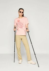 Jasnoróżowa koszulka z graficznym wzorem, beżowe spodnie i białe buty. Modelka korzysta z czarnych kijów trekkingowych i nosi białe okulary przeciwsłoneczne. Styl minimalistyczny.