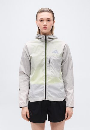 Młoda osoba stojąca, ubrana w jasnoszarą kurtkę z kapturem na zamek błyskawiczny z kieszenią na piersi oraz czarne szorty, na tle jednolitego białego tła.