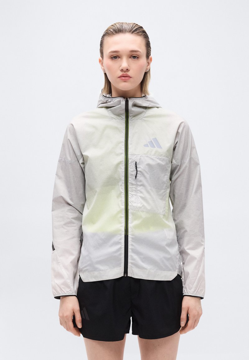 Νεαρό άτομο που στέκεται, φορά ανοιχτό γκρι φούτερ με κουκούλα και φερμουάρ, με τσέπη στο στήθος και μαύρα σορτς, μπροστά σε απλό λευκό φόντο.