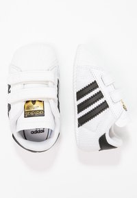 Vita adidas-sneakers med svarta ränder, med en texturerad ovandel, gummisula, kardborrestängning och en gulllogo på tungan.