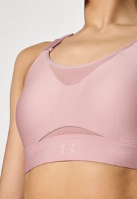 Lys rosa trenings-BH med teksturert overflate, rund hals og nettpanel. Under Armour-logo på den fremre stroppen. Justerbare stropper.