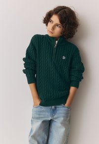 Jersey verde de punto con media cremallera, con puños y dobladillo acanalados. Se lleva con vaqueros de mezclilla de corte suelto en azul claro.