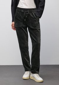 Persona che indossa pantaloni in velluto a costine neri con tasche frontali e sneakers bianche, in piedi contro uno sfondo grigio uniforme.