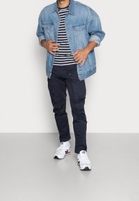 Kurtka dżinsowa, koszulka w paski, granatowe spodnie typu cargo oraz białe trampki. Kurtka posiada klasyczne kieszenie; koszulka ma poziome paski.