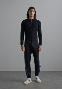 Tânăr cu părul până la umeri, îmbrăcat cu un polo negru cu mâneci lungi, pantaloni de trening negri cu șnur, având dungi pe laterale și încălțări sport albe.